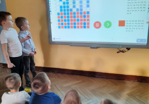 Dzieci siedzą na dywanie, a Oliwier i Krzyś stoją i próbują odgadnąć jaki napis pojawił się na tablicy multimedialnej.
