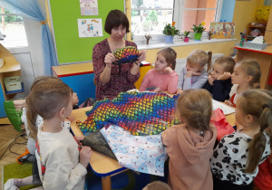 Dzieci przykucnęły wokół stołu, na którym leżą tkaniny, a Pani pokazuje dzieciom saszetkę uszytą z kolorowego materiału.