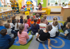 Dzieci siedzą na dywanie, a przed nimi siedzi przy stole mama Alicji. Pani prezentuje przedszkolakom maszynę do szycia i opowiada o zawodzie krawcowej.