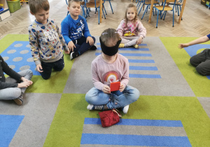 Dzieci z grupy "Słoneczka" siedzą na dywanie. Na środku siedzi Ala z zasłoniętymi oczami, losuje figurę geometryczną z woreczka.