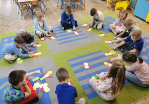 Dzieci siedzą na dywanie i układają przed sobą swój geometryczny obrazek z figur geometrycznych.