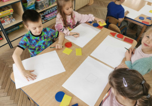 Krzysio, Vanessa i Ala siedzą przy stoliku i odrysowują na kartkach papieru figury geometryczne według instrukcji nauczyciela.