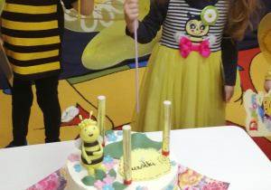Amanda i Mikołaj stoją z balonami, a przed nimi na białym stoliku stoi tort z pszczółką.