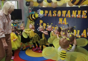 Nauczyciel prowadzi dzieci w przebraniach pszczółek. W tle dekoracja z okazji pasowania na przedszkolaka.
