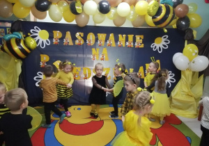 Dzieci tańczą w dwuosobowych kółeczkach. W tle dekoracja z okazji pasowania na przedszkolaka.