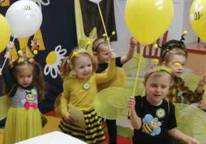 Grupka dzieci stoi w rozsypce trzymając balony żółte i białe oraz dyplomy. W tle dekoracja z okazji pasowania na przedszkolaka, a przed dziećmi stoi biały stoliczek.