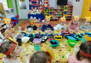 "Motylki" siedzą przy stole, na którym są pojemniki z ziemią, kolorowymi kamyczkami, "doniczkami", kolorowymi mazakami. W tle dekoracja: napis Dzień Misia, postacie pluszaków i misie przyniesione przez dzieci oraz kąciki zabaw.