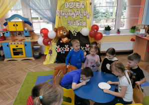 Oliwia i Krzyś siedzą przy stoliku podczas konkursu "Kto pierwszy zliże miód z talerzyka". Obok przykucnęło kilkoro dzieci. W tle dekoracja z napisem "Dzień Pluszowego Misia", balonami oraz misiami wykonanymi przez dzieci.
