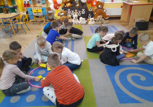 Dzieci siedzą na dywanie wokół trzech obręczy i układają misiowe puzzle. W tle fragment dekoracji: sylwety własnoręcznie wykonanych przez dzieci misiów przypięte do brązowego materiału, pluszaki siedzące na podłodze, misiowe balony.