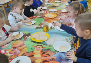 Dzieci siedzą przy złączonych stołach nakrytych ceratą z Kubusiem Puchatkiem i degustują miód na wafelkach.