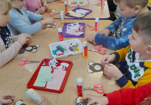 Dzieci siedzą przy złączonych stołach i naklejają elementy na sylwetę misia. Na stołach leżą pojemniki z kolorowymi kartkami, plasteliną, klej, nożyczki.