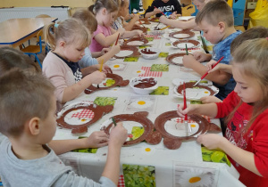 Dzieci siedzą przy stole i malują brązową farbą maskę misia z tekturowego talerzyka.