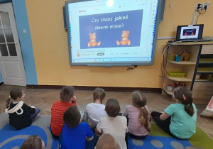 Dzieci siedzą na dywanie przed tablicą multimedialną i oglądają film "Dzień Pluszowego Misia".
