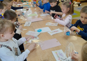 Dzieci siedzą przy stole i pakują do torebek foliowych swoje mydełka. Na stole leżą sznureczki, deski oraz papierowe torebki ozdobione przez dzieci.