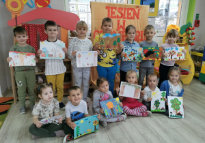 Dzieci, które wzięły udział w konkursie stoją na tle napisu i prezentują swoje prace konkursowe.