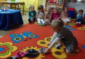 Oliwia dokłada swój bucik do innych butów ustawionych na dywanie. Wokół siedzą koledzy i koleżanki z grupy, w tle: kąciki zabaw i stolik do wróżb.