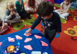 Filip wybiera jedną z różowych karteczek, na których są wypisane imiona dziewczynek. Karteczki leżą na niebieskim materiale, obok leżą karty do gry, papierowe duszki i klucze i kolorowy lampion. Z tyłu dzieci z grupy.