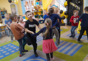 Dzieci tańczą w parach przy muzyce. Przy suficie wiszą balony w kolorze złotym i czarnym.