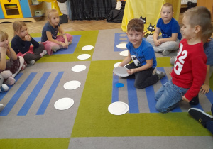Na dywanie leżą w dwóch rzędach białe talerzyki, a obok nich siedzą dzieci. Karol podniósł talerzyk i odkrył koło w kolorze niebieskim, co oznacza, że czekają go podróże. W tle dekoracja andrzejkowa.