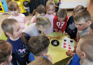 Dzieci stoją wokół stolika i oglądają wróżbę z wosku położoną na czarnym talerzyku. Obok leży klucz do lania wosku, dwie duże karty.