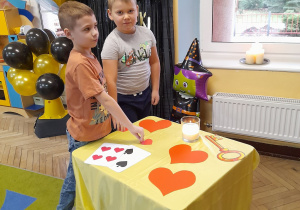 Dwóch chłopców stoi obok stolika i wybiera serduszko z imieniem dziewczynki. W tle dekoracja andrzejkowa.