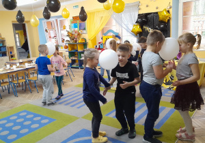 Dzieci tańczą na dywanie z balonami trzymając je między głowami. Przy suficie wiszą złote i czarne balony.