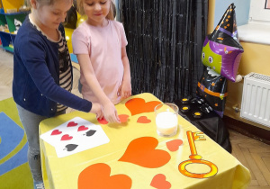 Alicja i Vanessa stoją przy stoliku nakrytym żółtym materiałem, na którym leżą serca wycięte z kartonu, karty, klucz andrzejkowy, a obok stoi świeca. Dziewczynki wybierają serce z imieniem chłopca. W tle dekoracja andrzejkowa.