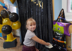 Alicja uklęknęła przed dekoracją andrzejkową. Dziewczynka wyjmuje z magicznego woreczka położonego obok balonowej wróżki kartę z zawodem, jaki będzie wykonywać w przyszłości.