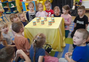 Dzieci przykucnęły wokół stolika nakrytego żółtym materiałem, na którym stoją kubki z wróżbą. W tle dekoracja andrzejkowa, kącik domowy, przyrodniczy, okno.