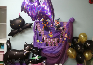 Dekoracja z okazji Andrzejek: na tablicy pokrytej fioletowym materiałem znajduje się napis Andrzejki, kolorowe czarownice oraz płonąca świeca. Z boku stoją w wazonie balony w kolorze złotym i czarnym. Z drugiej strony na szafce pokrytej fioletowym materiałem stoi czarny balonowy kot, nad którym wisi czarny oraz fioletowy balonowy nietoperz, obok szafki stoi balonowa głowa czarownicy. Przed tablicą stoi stoliczek pokryty fioletowym materiałem na którym leży klucz, karty do gry, dwa czerwone serca, oraz zapalone świeczki w latarenkach.