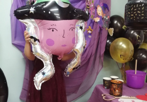 Tosia pozuje do zdjęcia z balonową głową czarownicy na tle dekoracji.