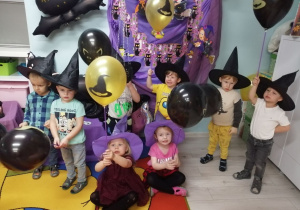 Zdjęcie grupowe dzieci na tle dekoracji. Przedszkolaki w czarnych kapeluszach trzymają balony w kolorze złotym i czarnym.