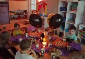 Dzieci siedzą przy wspólnym stole i degustują słodki poczęstunek. Na stole stoją w wazonie czarne balony , słodycze oraz zapalone lampiony, stół przykryty jest fioletowym obrusem.