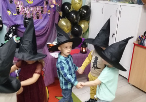 Dzieci tańczą w małych kółeczkach. Dzieci mają na głowie założone czarne kapelusze czarownicy. W tle dekoracja z okazji Andrzejek.
