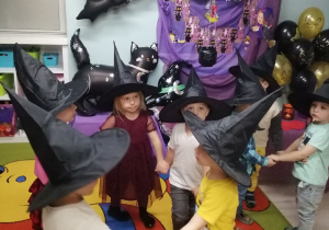„Pszczółki” trzymają się za ręce w kółeczku. Dzieci mają na głowie założone czarne kapelusze czarownicy. W tle dekoracja z okazji Andrzejek.
