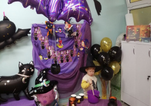 Bruno w czarnym kapeluszu miesza w kociołku. Kociołek stoi na stoliczku pokrytym fioletowym materiałem.W tle dekoracja z okazji Andrzejek.