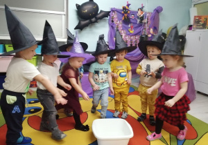Dzieci stoją w półkolu i wrzucają do białej miski pieniążki. Przedszkolaki mają na głowach założone czarne kapelusze czarownicy.