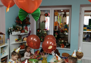 Dzieci siedzą przy złączonych stołach nakrytych ceratą z Kubusiem Puchatkiem i kolorują mazakami sylwetę misia. Na środku stołu w wazonie stoją brązowe misiowe balony, oraz na suficie wiszą balonowe głowy misia z kolorowymi muchami.