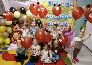 Zdjęcie grupowe dzieci z balonami w kształcie misia na tle dekoracji. Dzieci mają na głowach papierowe misiowe czapeczki.