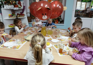 Dzieci siedzą przy złączonych stołach na których leżą białe serwetki w misie i degustują miód na wafelkach. Na środku stołu w wazonie stoją brązowe misiowe balony,lemoniada w dzbanku i słoiczek miodu.