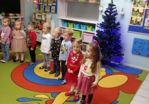 Dzieci ustawione w półkolu śpiewają piosenkę. W tle choinka ubrana w świecące niebieskie lampki , wiszące reniferki wykonane przez dzieci z szarych torebek oraz półki z klockami.