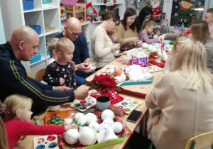 Dzieci wraz z rodzicami siedzą przy wspólnym stole i ozdabiają styropianowe kule .Na stołach pokrytych świątecznymi serwetkami leżą na kolorowych tackach styropianowe kule, kolorowe tasiemki, cekiny.