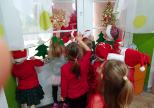 Motylki stoją przy oknie, przez które zagląda Mikołaj w czerwonym ubraniu i z białą brodą.