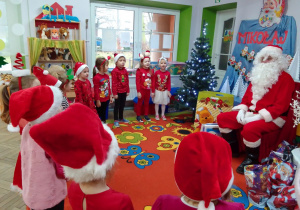 Dzieci w mikołajowych czapkach stoją w półkolu i prezentują piosenkę „Grudzień z Mikołajem”. Na wprost Motylków siedzi Święty Mikołaj, w tle dekoracja: napis Mikołaj, girlanda z Mikołajami, choinka z lampkami.
