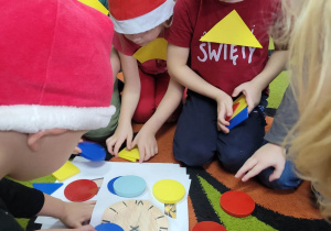 Biedronki zgromadzone na dywanie wokół fotografii przedstawiającej zegar. Dzieci odgadują jaki figurę geometryczną widzą na zdjęciu i dokładają figury kół