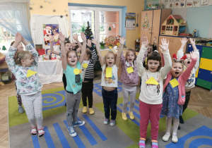 Dzieci z grupy "Słoneczka" stpja na dywanie z podniesionymi rękoma w górze podczas zabawy przy muzyce "Powitanie z okrzykiem".