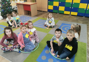 Dzieci z grupy "Słoneczka" siedzą odpowiednio w oznaczonych obręczach. Każde z dzieci ma przyczepioną do bluzki jedną figurę geometryczną: koło, kwadrat, trójkąt lub prostokąt.