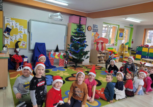 Dzieci z grupy Biedronek siedzą na dywanie w oczekiwaniu na Świętego Mikołaja. Każde dziecko ma założoną mikołajkową czapkę lub opaskę z wizerunkiem renifera. W tle widać świąteczną dekorację w sali.