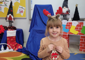 Zuzia pozuje do zdjęcia na tle świątecznej dekoracji. Dziewczynka ma na głowie opaskę przedstawiającą renifera, a w rączkach trzyma bombkę z wygrawerowanym swoim imieniem.