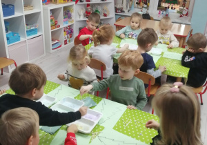 Dzieci siedzą przy stolikach pokrytych zieloną ceratką i wykonują pracę plastyczną: stemplują patyczkami na konturze choinki.